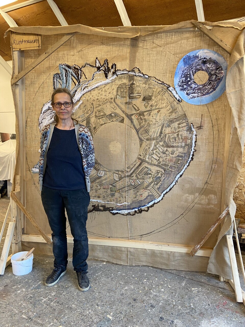 Kunstner Marianne Jørgensen foran sit broderi af Nuuk, formet som en kæmpeperle med hul i midten. I øverste højre hjørne ses fotografiet af Nuuk taget fra en af de højeste kraner i hovedstaden.